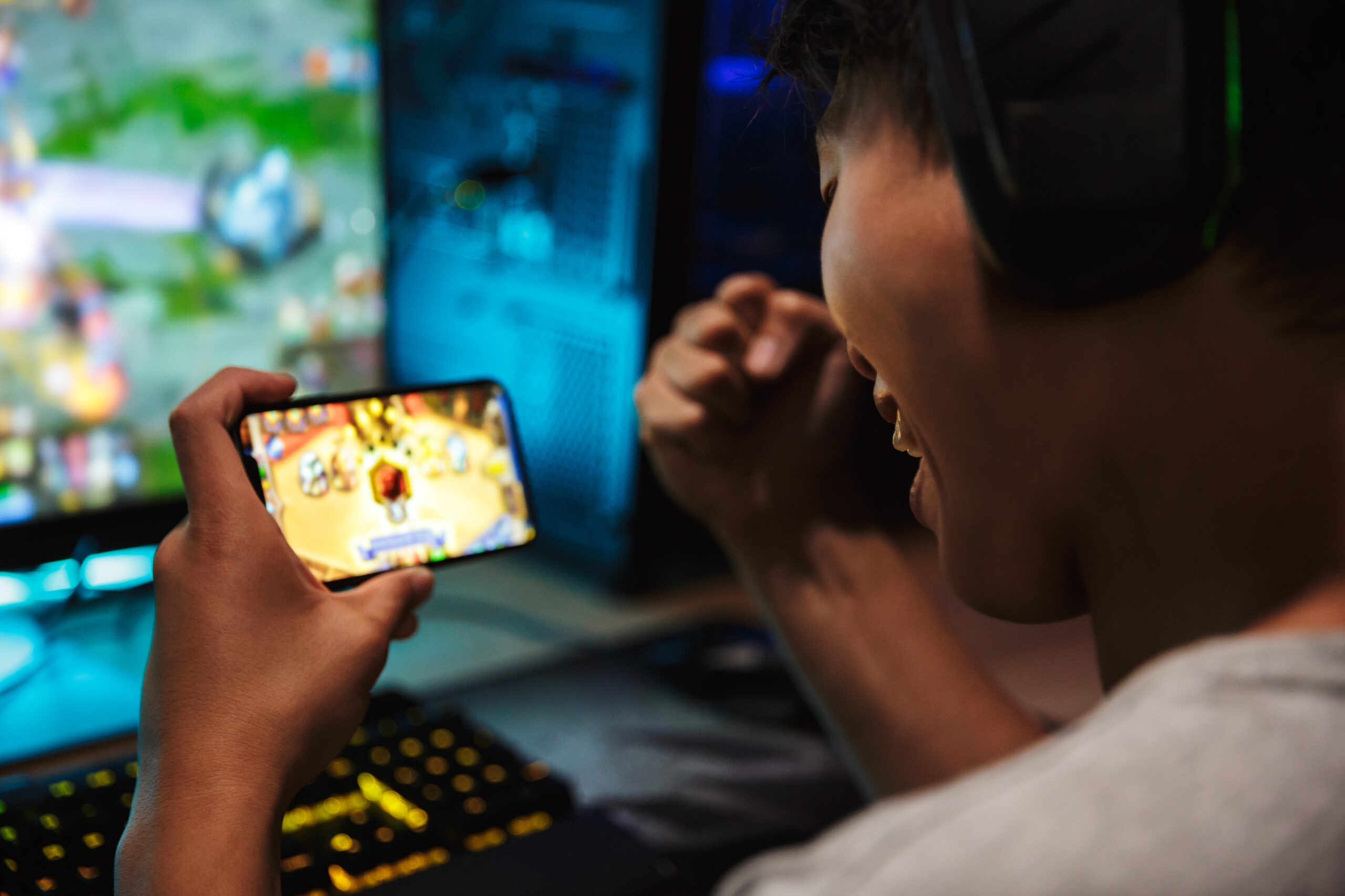 Image of teenage gamer boy playing video games on smartphone and computer in dark room wearing headphones and using backlit rainbow keyboard